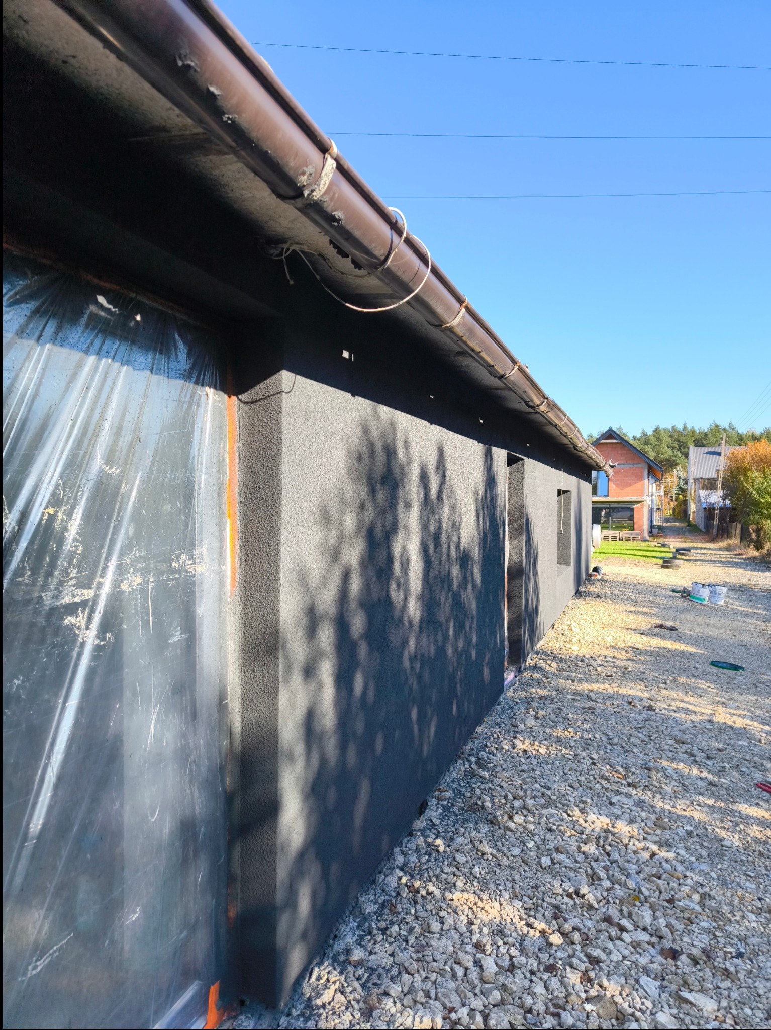 Szara elewacja budynku w trakcie ocieplania, zabezpieczona folią, widoczne brązowe rynny i kamieniste podłoże.