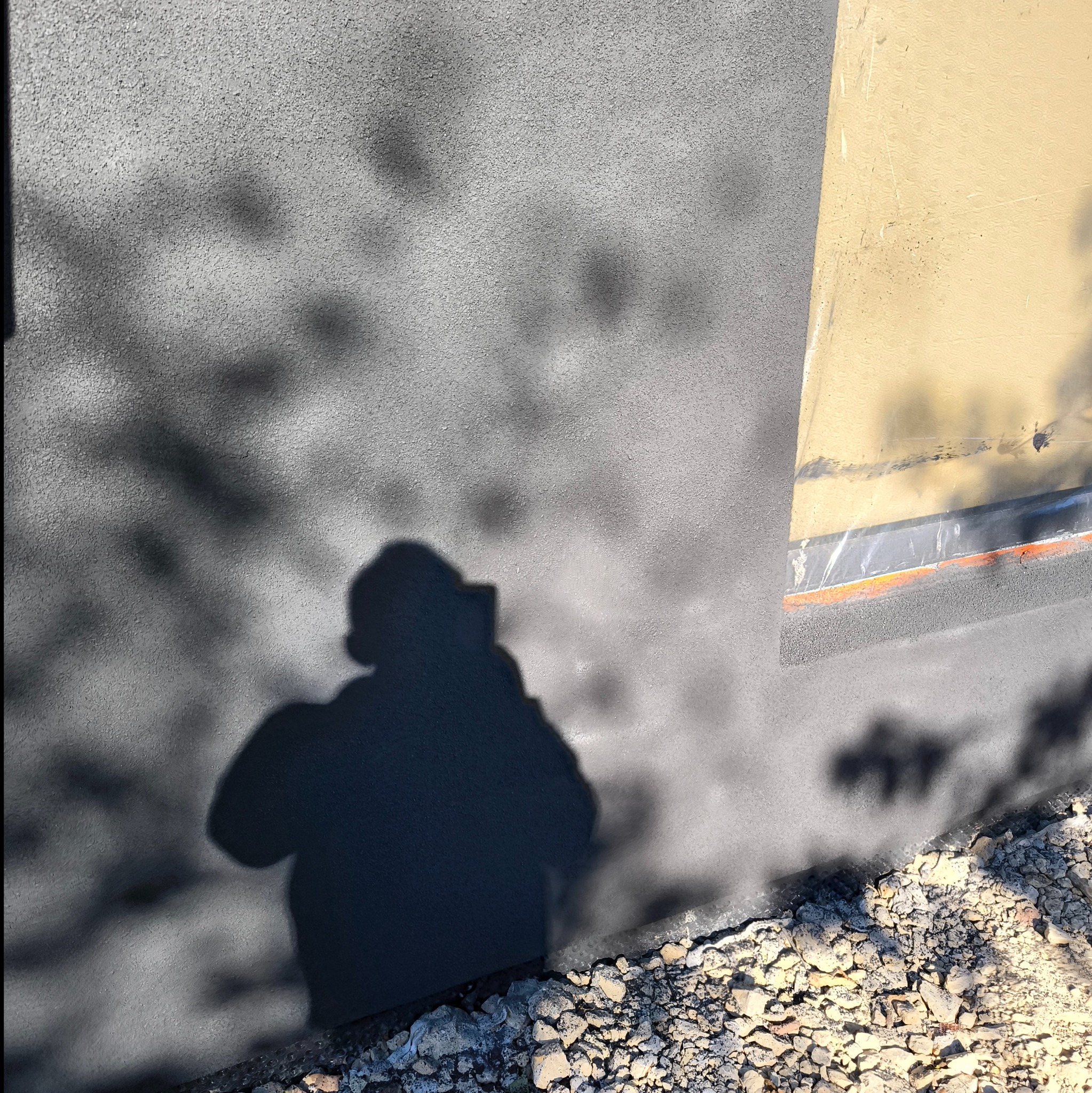Szara elewacja z widocznym cieniem osoby robiącej zdjęcie, obok fragment okna zabezpieczonego folią, u dołu kamienie.