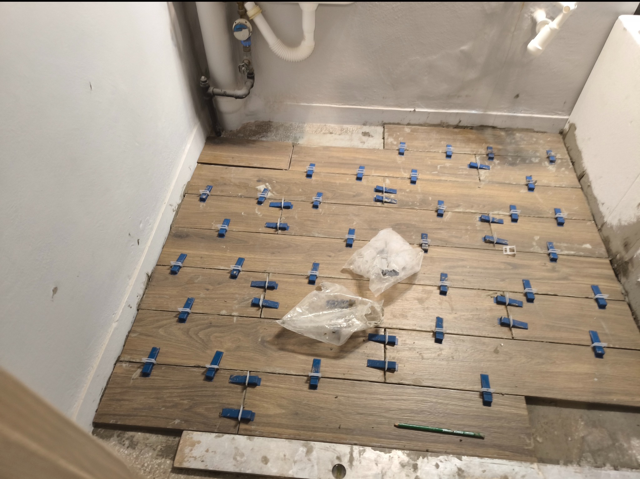 Podłoga w trakcie układania płytek imitujących drewno w łazience, z zastosowaniem niebieskich klipsów poziomujących; widoczne worki z klejem i ołówek na podłodze.