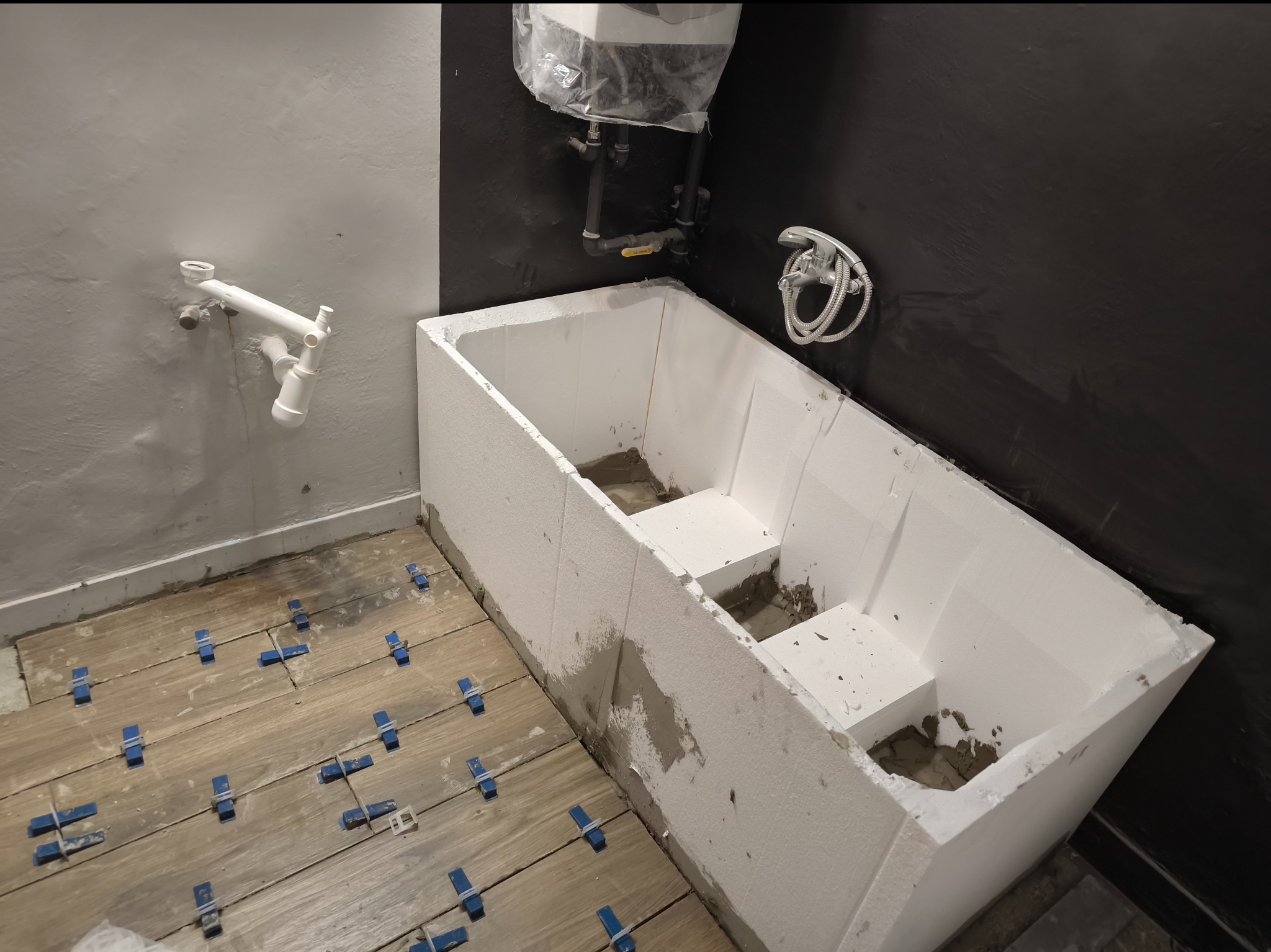 Wnętrze łazienki w trakcie remontu: zabudowa wanny z białych płyt, drewnopodobne płytki podłogowe z niebieskimi krzyżykami dystansowymi, rury odpływowe na ścianie i bateria prysznicowa na czarnej...