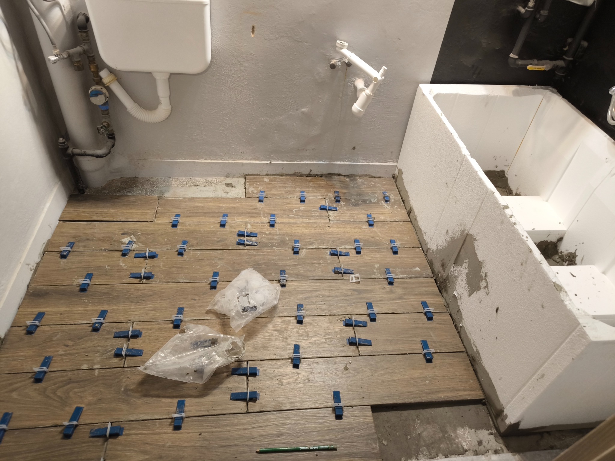 Ułożone płytki podłogowe imitujące drewno, z niebieskimi krzyżykami dystansowymi, widoczne worki foliowe, ołówek na podłodze, fragment obudowy wanny z białego styropianu, instalacja hydrauliczna...