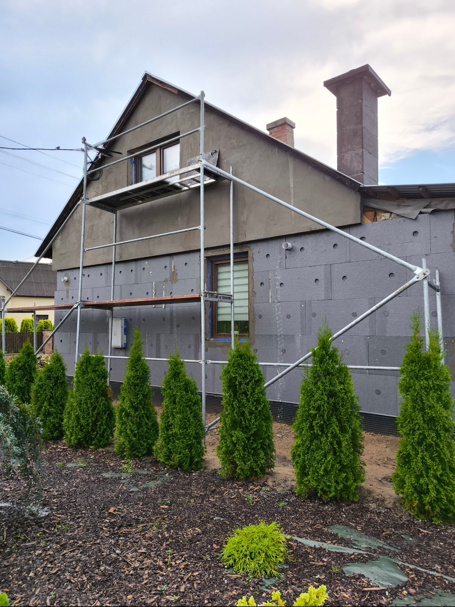 Dom jednorodzinny w trakcie ocieplania elewacji styropianem grafitowym, widoczne rusztowanie, fragment elewacji już otynkowany, rząd tuj szmaragdowych wzdłuż ściany budynku.