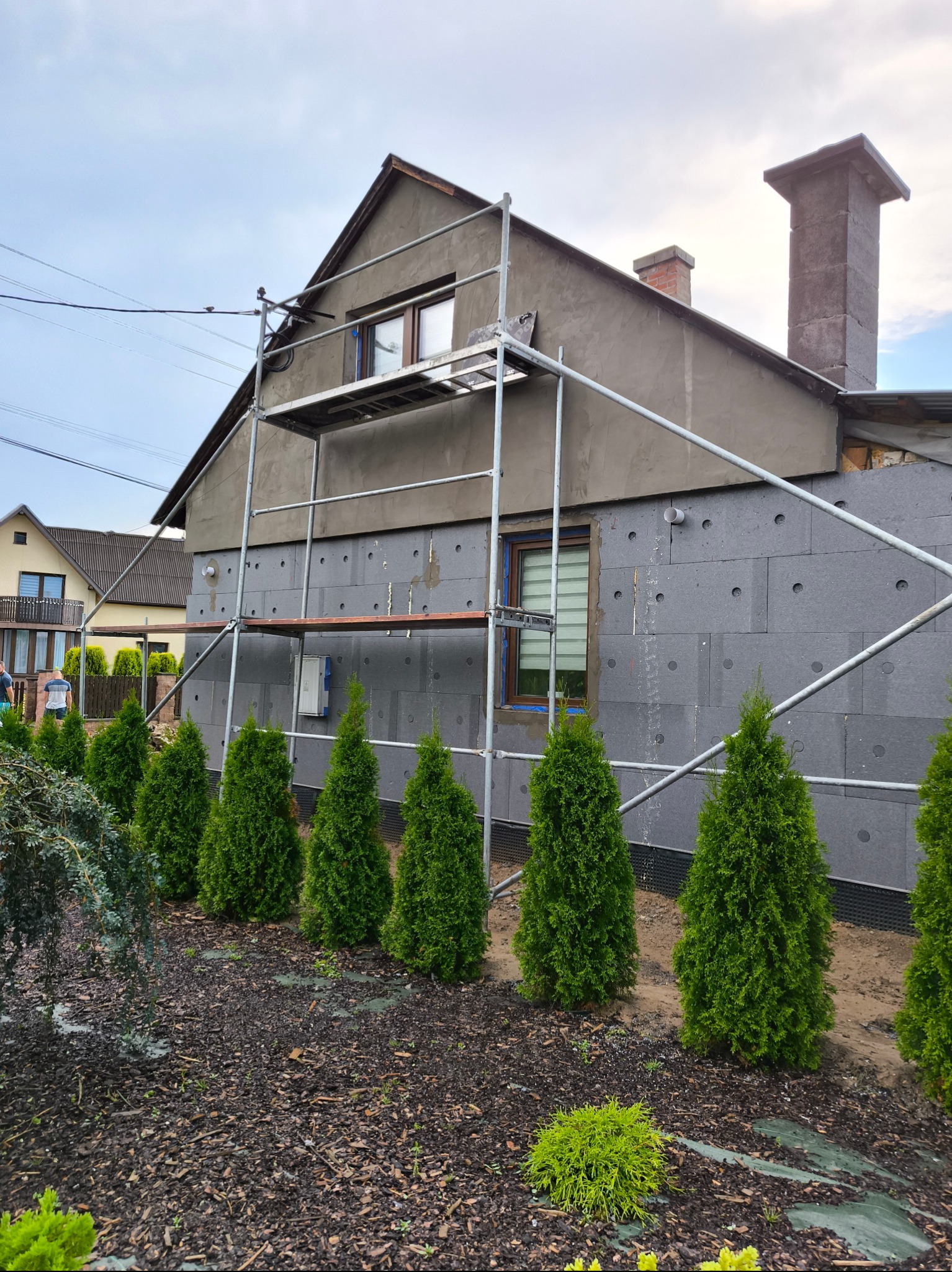 Ocieplanie elewacji budynku styropianem w Jerzmanowicach, widoczne rusztowanie, rząd małych tui i fragment sąsiedniego domu.