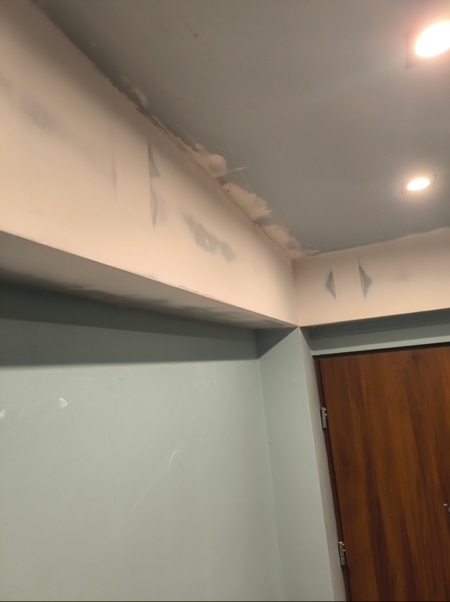 Fragment wnętrza w trakcie prac wykończeniowych: ściany i sufit po szpachlowaniu, widoczne ślady po wałku malarskim i niedoskonałości w narożnikach, przygotowane do malowania.