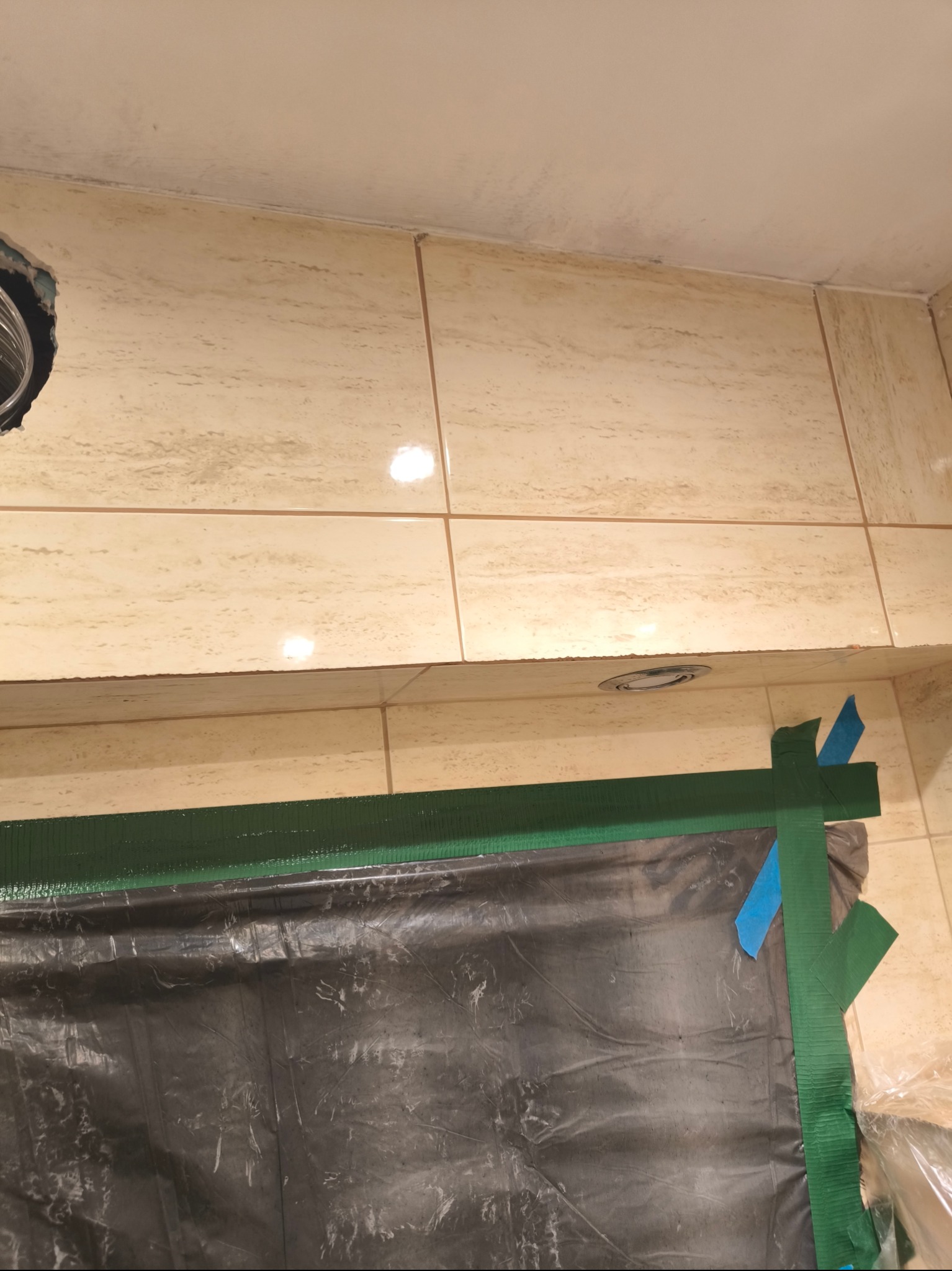 Przygotowanie do prac wykończeniowych: Zabezpieczona folią ściana z beżowych płytek, oklejona zieloną taśmą malarską i niebieskimi paskami. Widoczny otwór wentylacyjny w suficie i zamaskowane...