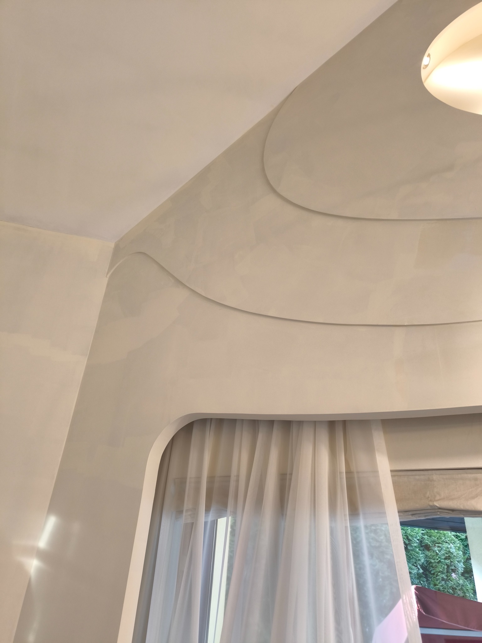 Nowoczesne wnętrze z dekoracyjną ścianą w odcieniach bieli, z subtelnymi, falistymi wgłębieniami pod sufitem i oknem zasłoniętym przezroczystymi firanami, oświetlone lampą sufitową