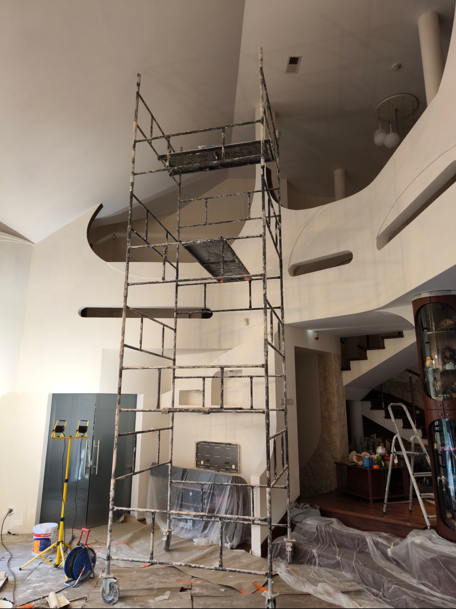 Wysoki, metalowy ruszt ustawiony w przestronnym salonie z antresolą, przygotowanym do prac wykończeniowych; podłoga i meble zabezpieczone folią, widoczne lampy budowlane i wiadro z farbą.