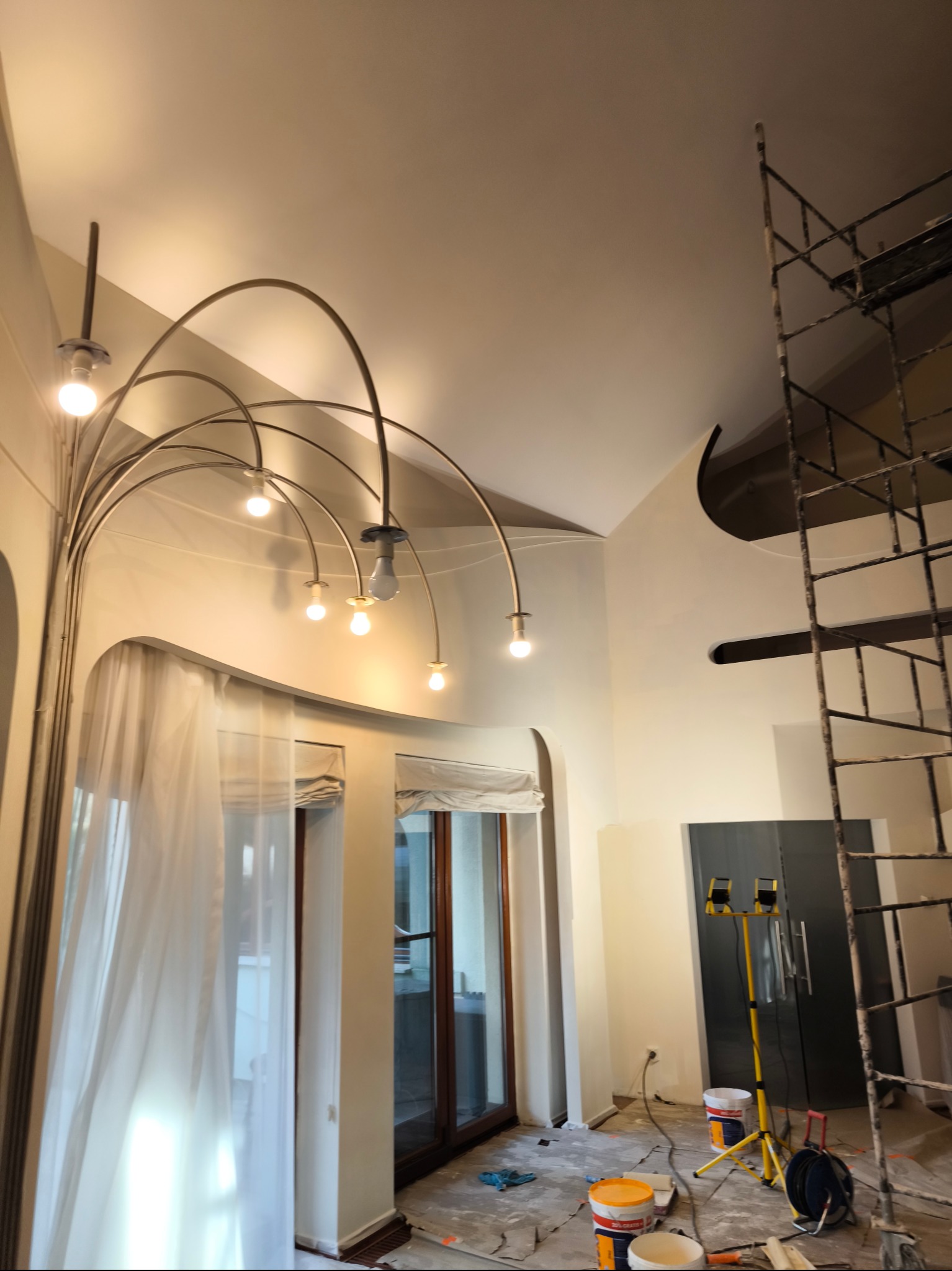 Wnętrze w trakcie prac wykończeniowych: widoczne rusztowanie, lampa budowlana na statywie, wiaderka z farbą i charakterystyczna lampa sufitowa z łukowatymi ramionami i żarówkami, ślady po malowaniu...