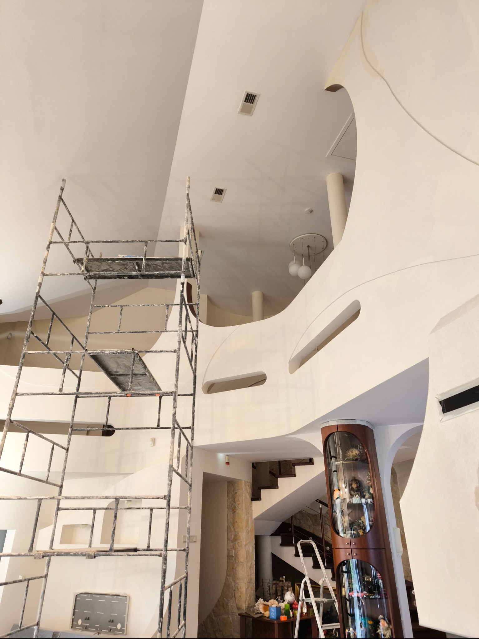 Wnętrze domu w trakcie remontu, widoczne rusztowanie, obudowa schodów z zaokrąglonymi ścianami w kolorze kremowym, częściowo wyszpachlowane ściany, na podłodze widoczne narzędzia i materiały...