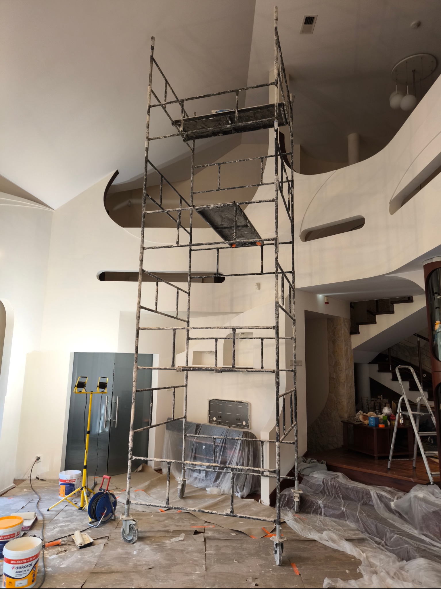 Wnętrze w trakcie odświeżania: wysoki, metalowy ruszt na kółkach stoi na zabezpieczonej folią podłodze, widoczne puszki z farbą, wałki malarskie, lampa budowlana, w tle ściany z zaokrąglonymi...