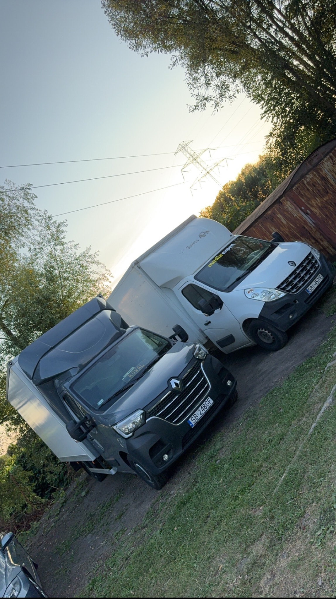 Dwa samochody dostawcze Renault Master, jeden szary i jeden biały, zaparkowane na trawiastym terenie w pobliżu metalowego ogrodzenia, z widocznymi liniami wysokiego napięcia i drzewami w tle...