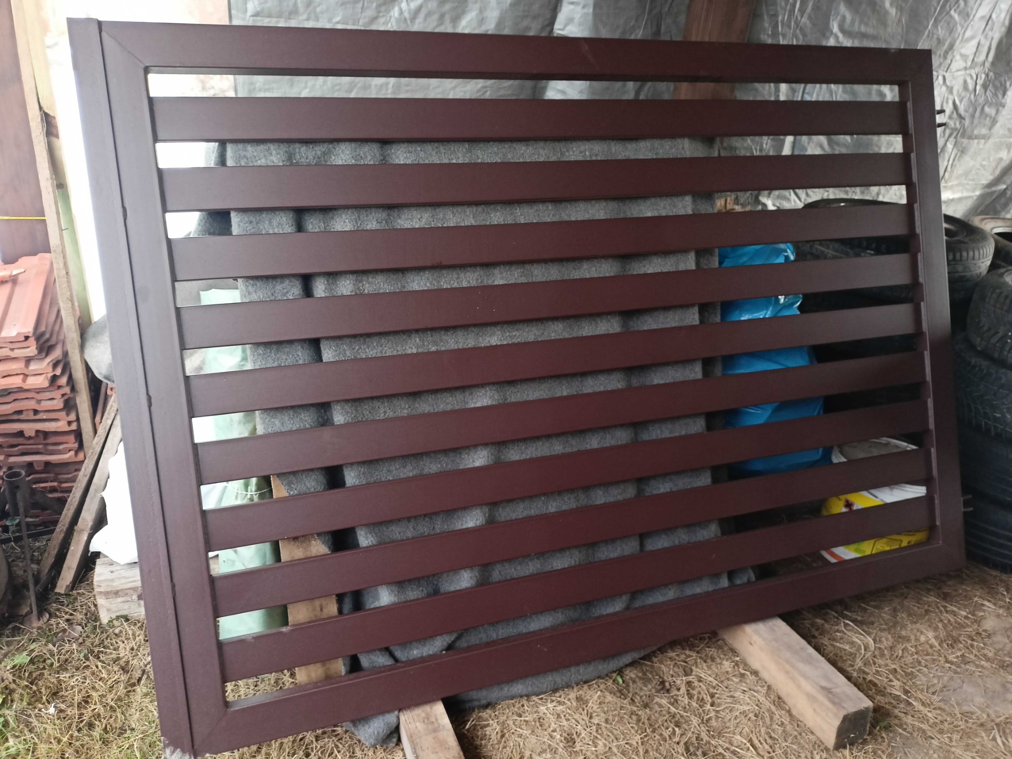 Gotowy, brązowy panel ogrodzeniowy z poziomymi szczeblami, oparty o drewniane belki na zewnątrz. Widoczne opony i dachówki w tle.