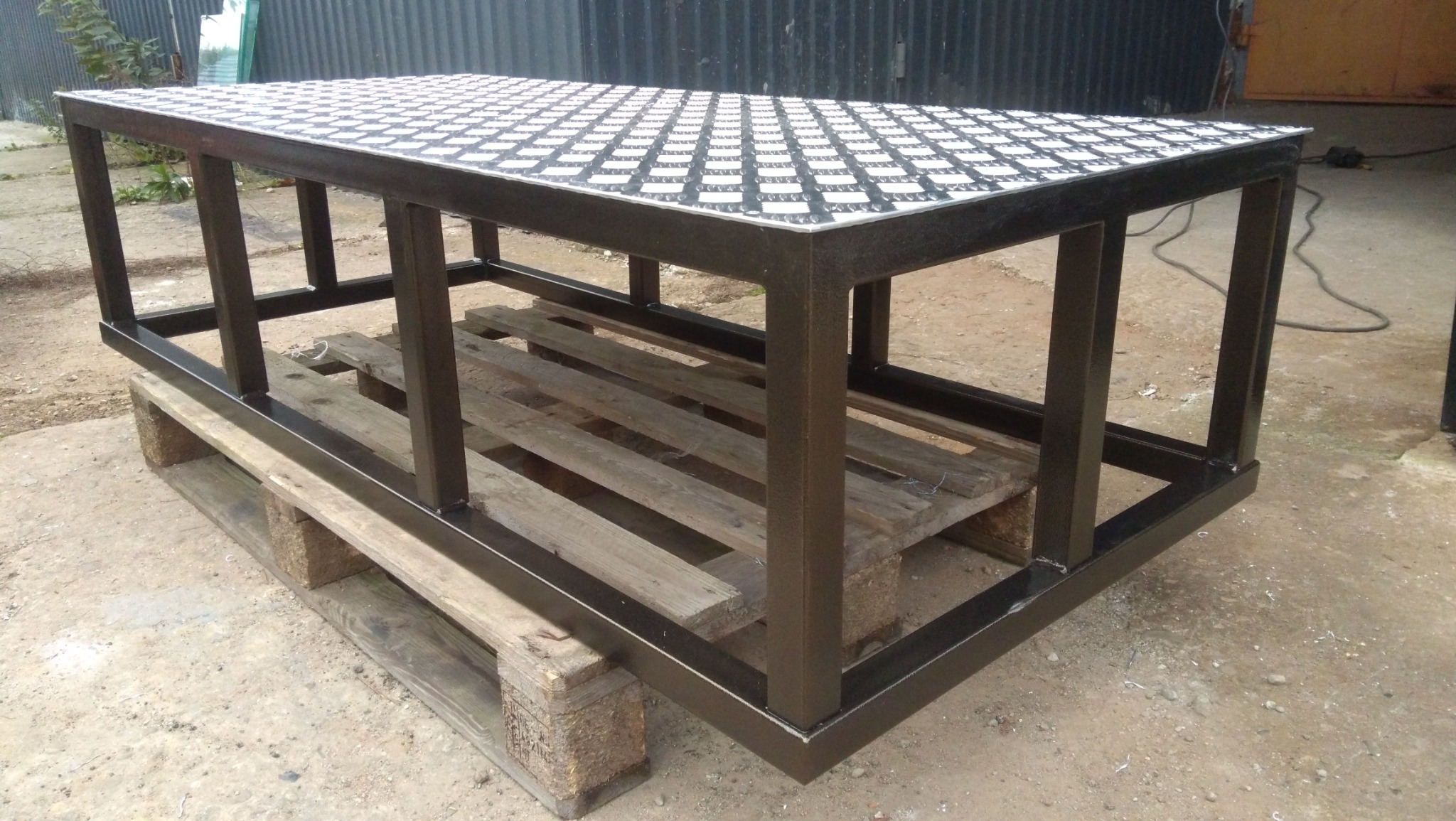 Metalowa konstrukcja stołu z blatem w szachownicę, spawana z profili, stojąca na paletach. Tło: blaszana ściana. Widok z boku, lekko z góry.