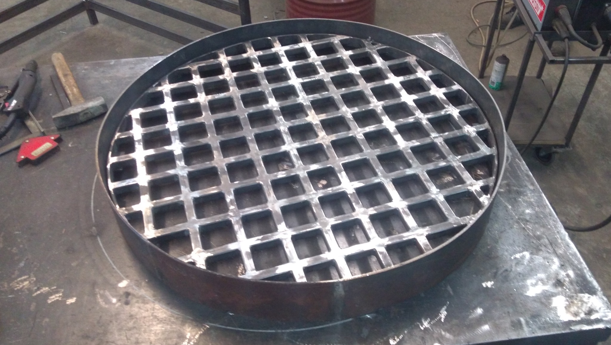 Okrągła metalowa konstrukcja z kratownicą, otoczona grubym pierścieniem, leży na metalowym stole warsztatowym. Obok widoczne narzędzia, w tle wózek spawalniczy.