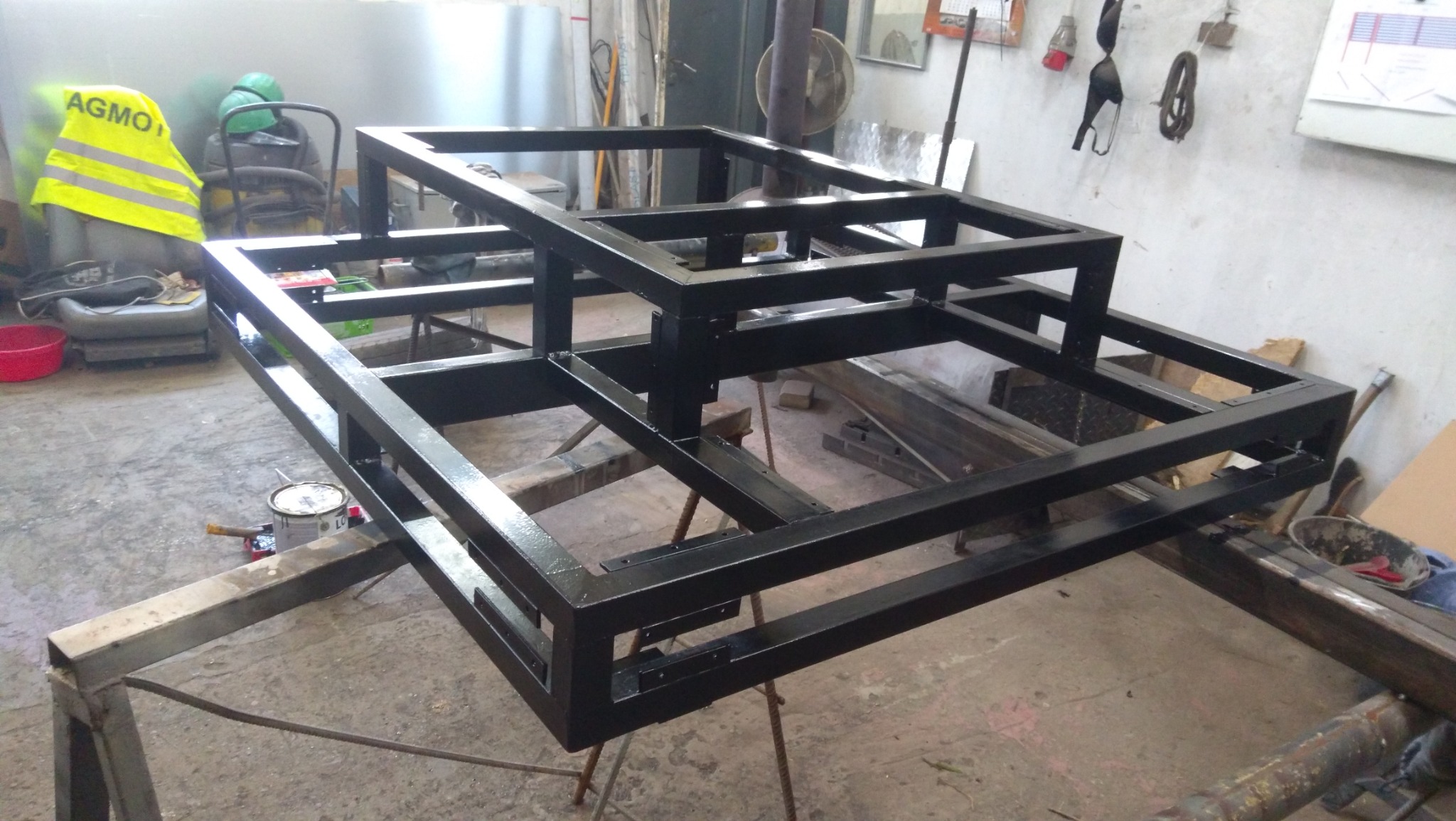 Czarna, metalowa konstrukcja szkieletowa na warsztacie, świeżo po malowaniu. Widoczny pojemnik z farbą, odzież robocza i narzędzia. Surowe, industrialne otoczenie.