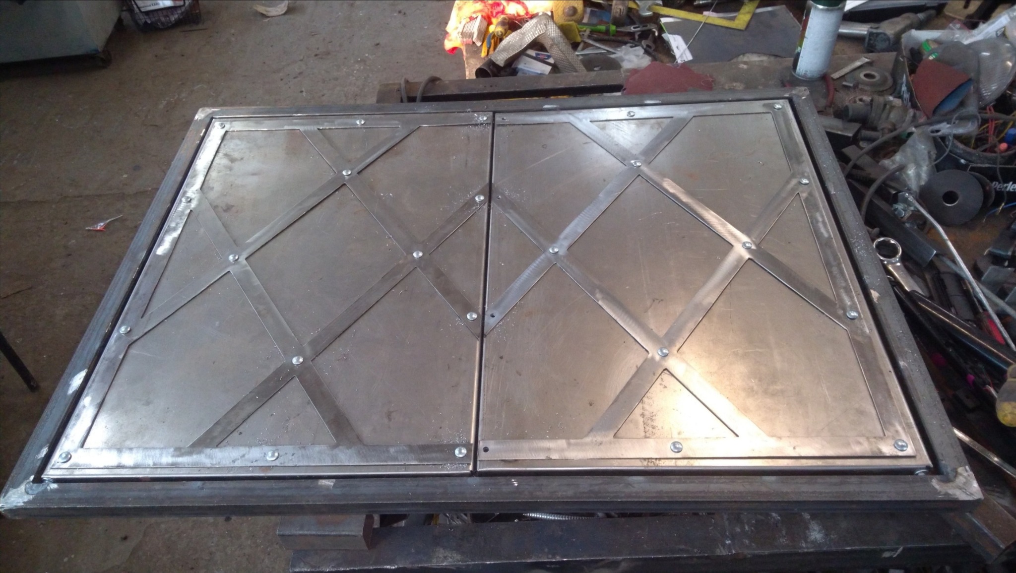 Metalowa, spawana konstrukcja z dwoma kwadratowymi panelami, wzmocniona ukośnymi listwami i nitami, leżąca na warsztatowym stole w otoczeniu narzędzi.