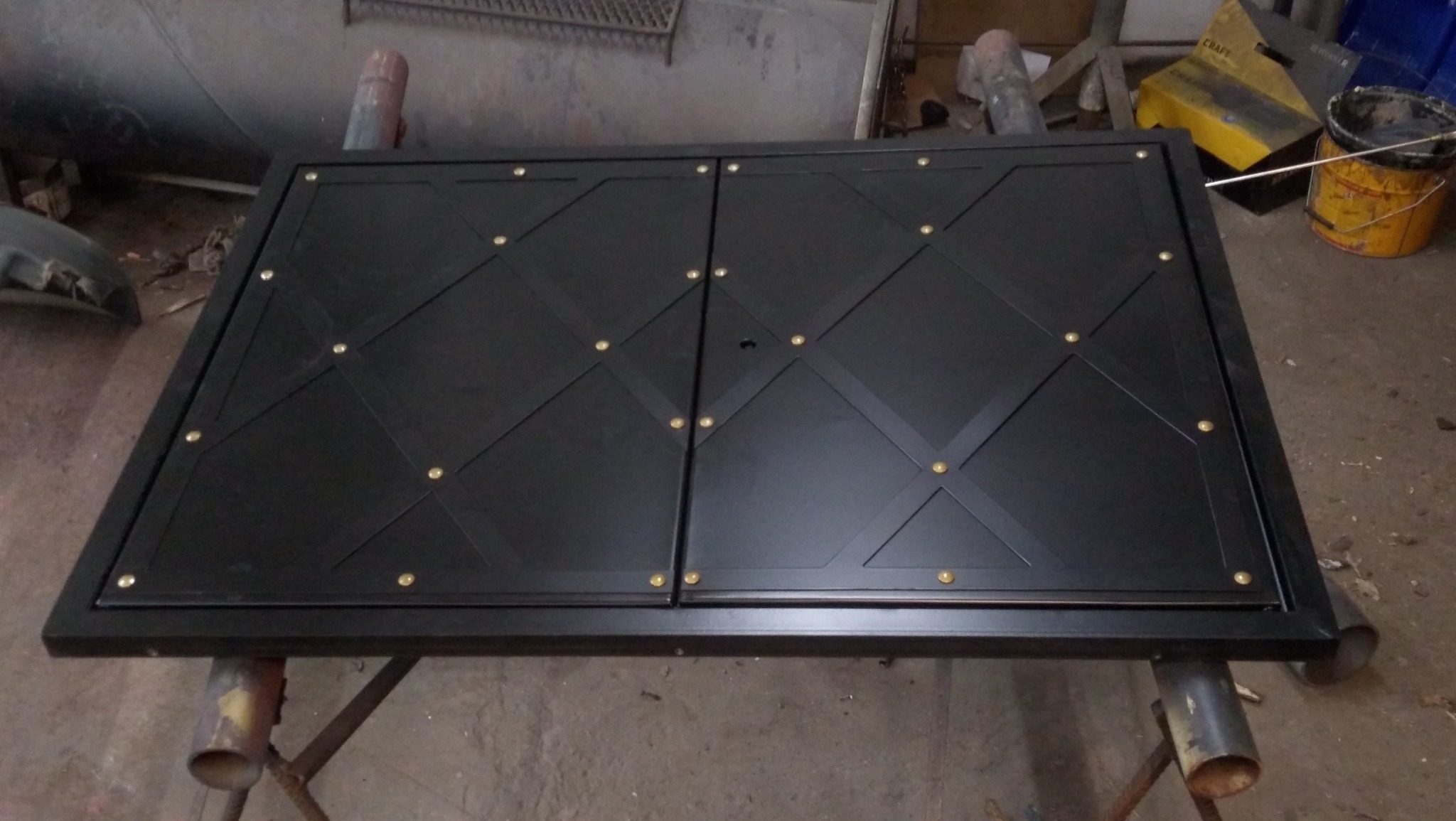 Czarna, metalowa brama dwuskrzydłowa z dekoracyjnymi, złotymi nitami, leżąca na metalowym stole w warsztacie, gotowa do montażu.