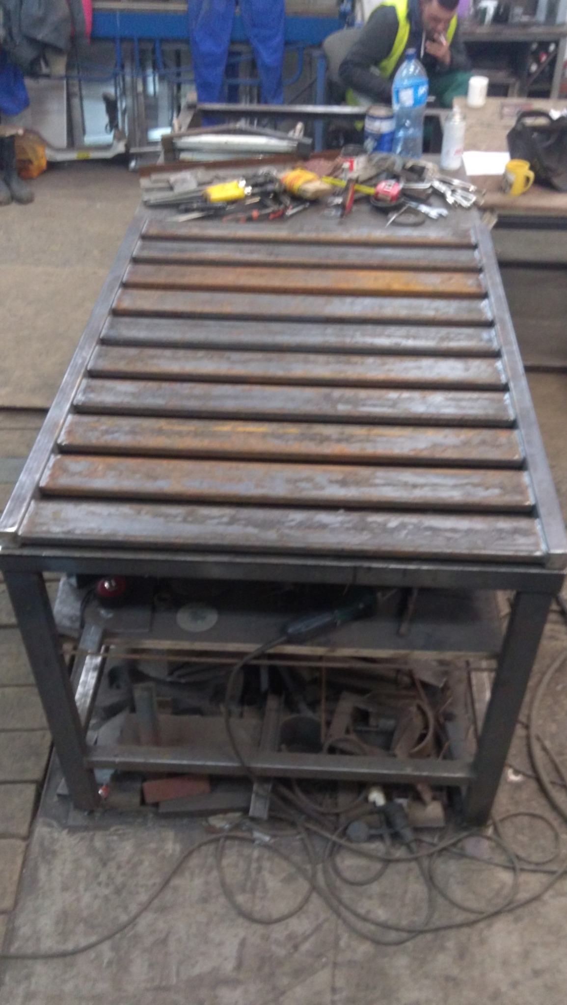 Metalowa konstrukcja stołu spawalniczego z narzędziami na blacie, w warsztacie. Widoczny spawacz w tle. Surowy wygląd, metalowe elementy, narzędzia pracy.