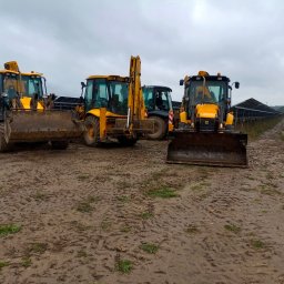 Domar - Trzy żółte koparko-ładowarki JCB zaparkowane na błotnistym terenie obok rzędu paneli słonecznych w pochmurny dzień.