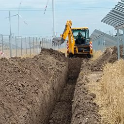 Domar - Żółta koparka JCB wykopuje wąski rów w ziemi na tle farmy wiatrowej i paneli słonecznych.