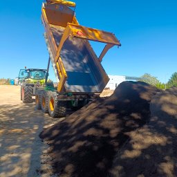 Domar - Żółta wywrotka z naczepą na tle błękitnego nieba, wysypująca ciemną ziemię, obok zielony traktor CLAAS i budynek w oddali.