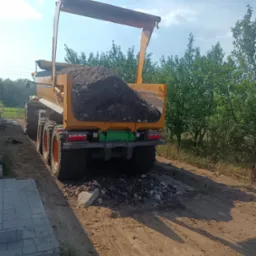 Żółta wywrotka z ładunkiem ciemnej ziemi lub gruzu na nierównej, piaszczystej drodze w słoneczny dzień, widok z tyłu.