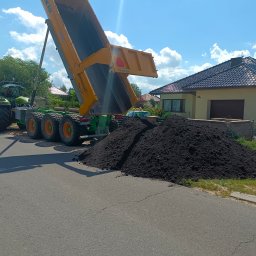 Domar - Zielony traktor z żółtą przyczepą wyładowuje ciemną ziemię na poboczu asfaltowej drogi, w tle dom jednorodzinny pod błękitnym niebem.