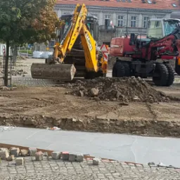 Przygotowanie terenu pod budowę: żółta koparka JCB i czerwona Liebherr na placu budowy, widoczny wykop z betonową wylewką i porozrzucane kostki brukowe, w tle budynki miejskie.
