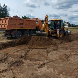 Domar - Żółta koparko-ładowarka JCB i pomarańczowa wywrotka podczas prac ziemnych na placu budowy, widoczne ślady opon w ziemi i wykop pod szambo.