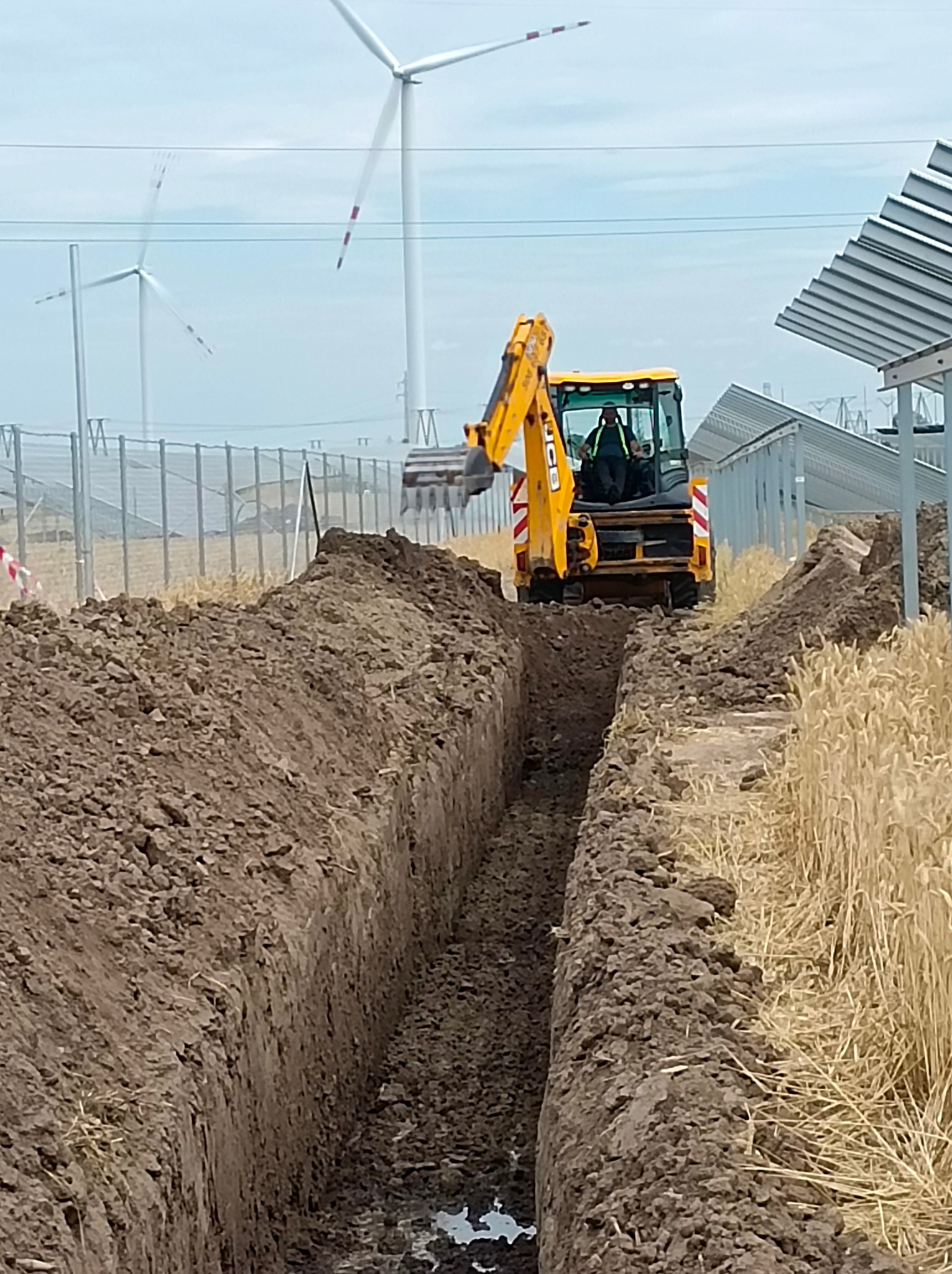 Żółta koparka JCB wykopuje wąski rów w ziemi na tle farmy wiatrowej i paneli słonecznych.