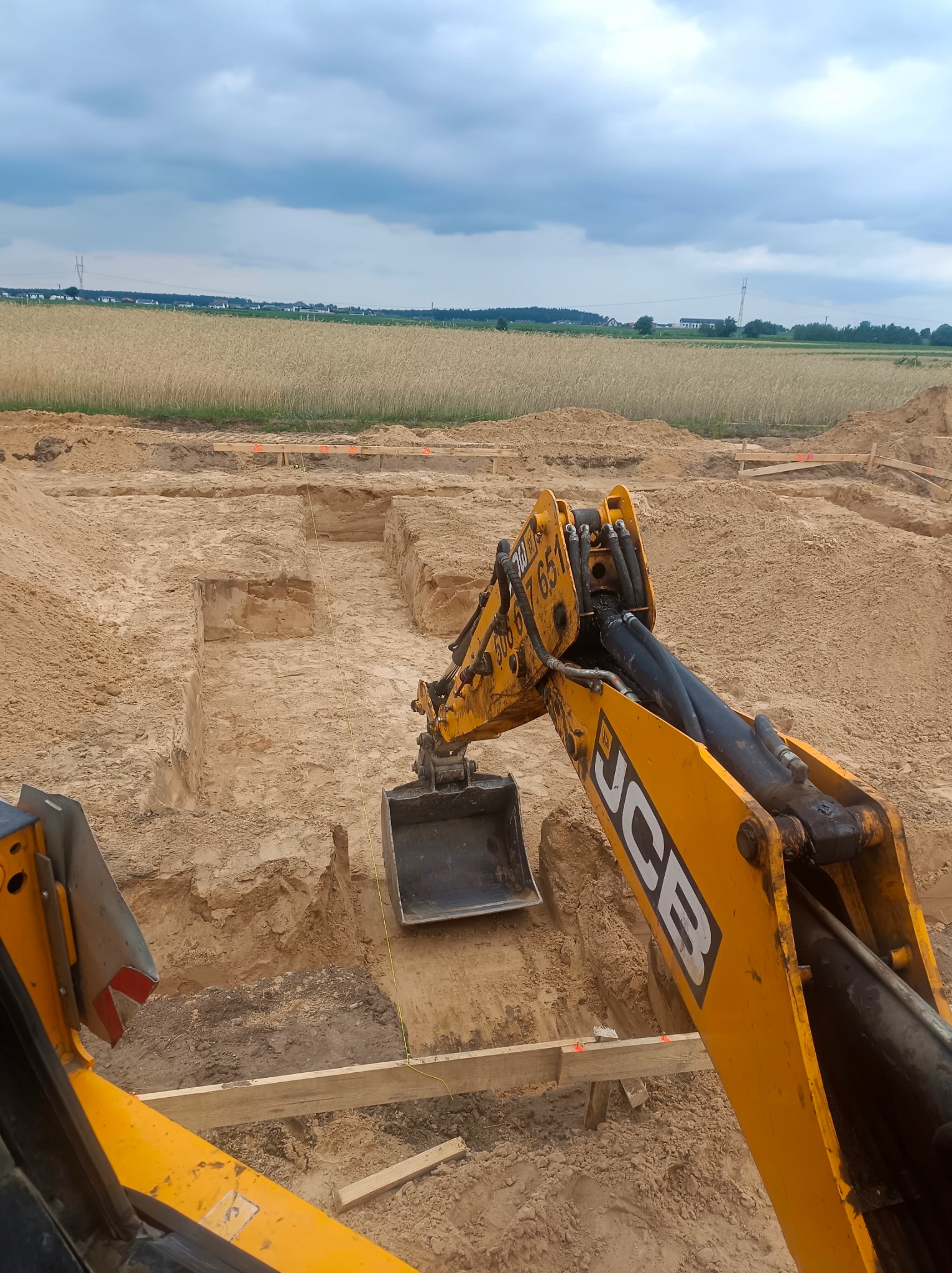 Żółta koparka JCB w trakcie prac ziemnych, widok z kabiny operatora. Wykopane fundamenty pod budynek, w tle pole uprawne pod pochmurnym niebem.
