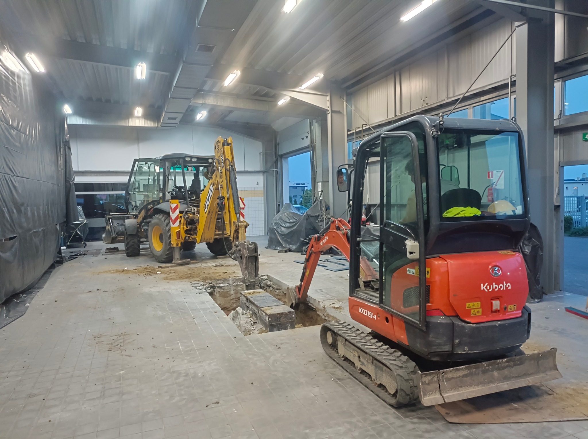 Wnętrze hali z dwoma maszynami budowlanymi: żółtą koparko-ładowarką JCB i pomarańczową mini koparką Kubota KX019-4, prowadzącymi prace ziemne wewnątrz budynku.