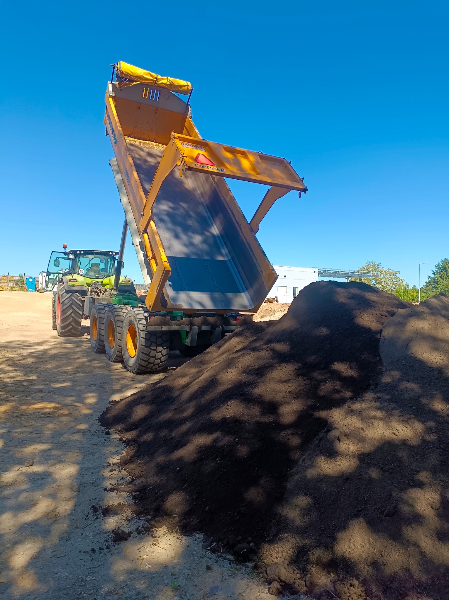Żółta wywrotka z naczepą na tle błękitnego nieba, wysypująca ciemną ziemię, obok zielony traktor CLAAS i budynek w oddali.