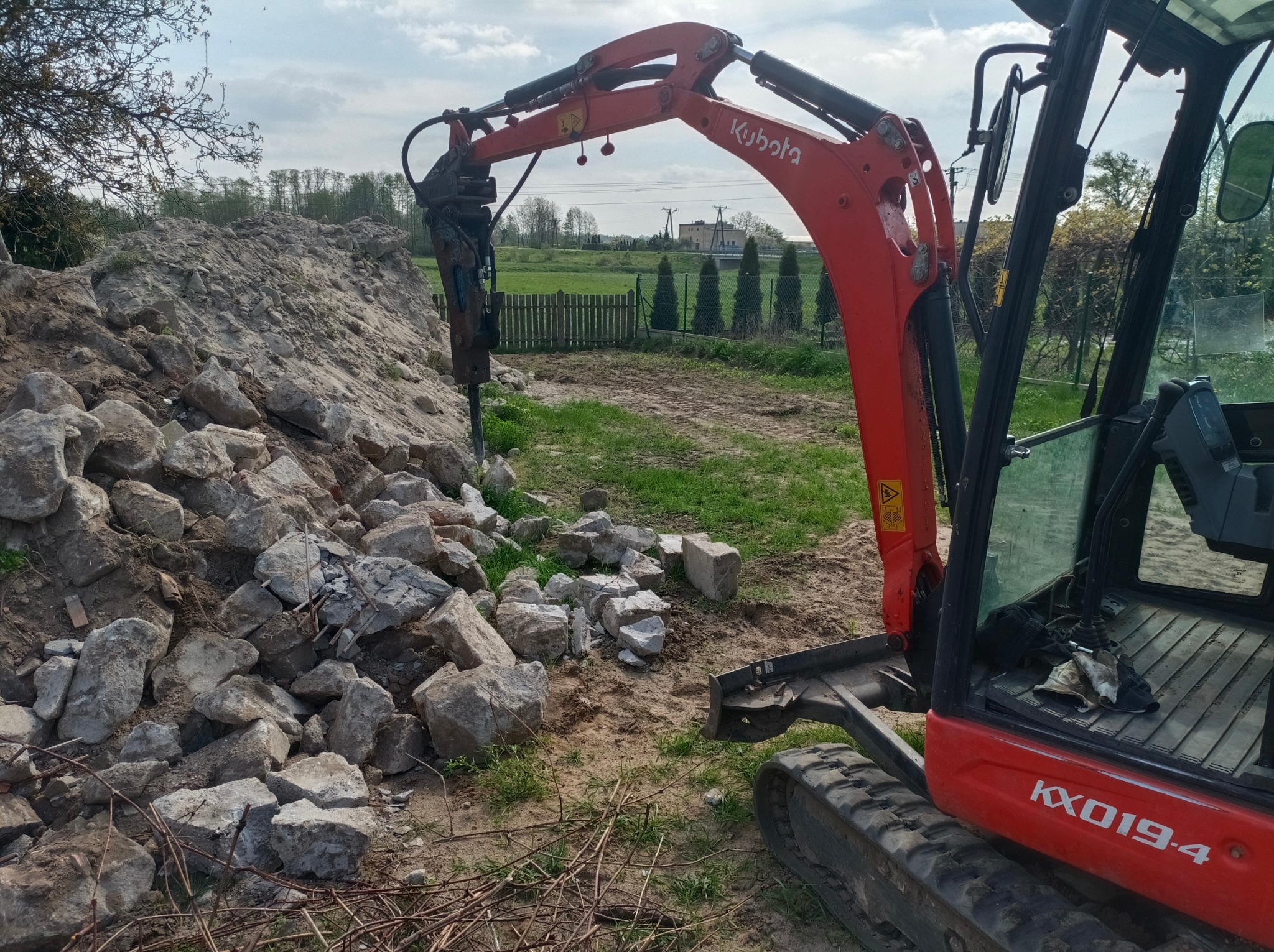 Czerwona minikoparka Kubota KX019-4 z młotem hydraulicznym kruszy skały i ziemię na placu budowy, w tle zielone pola i drzewa.