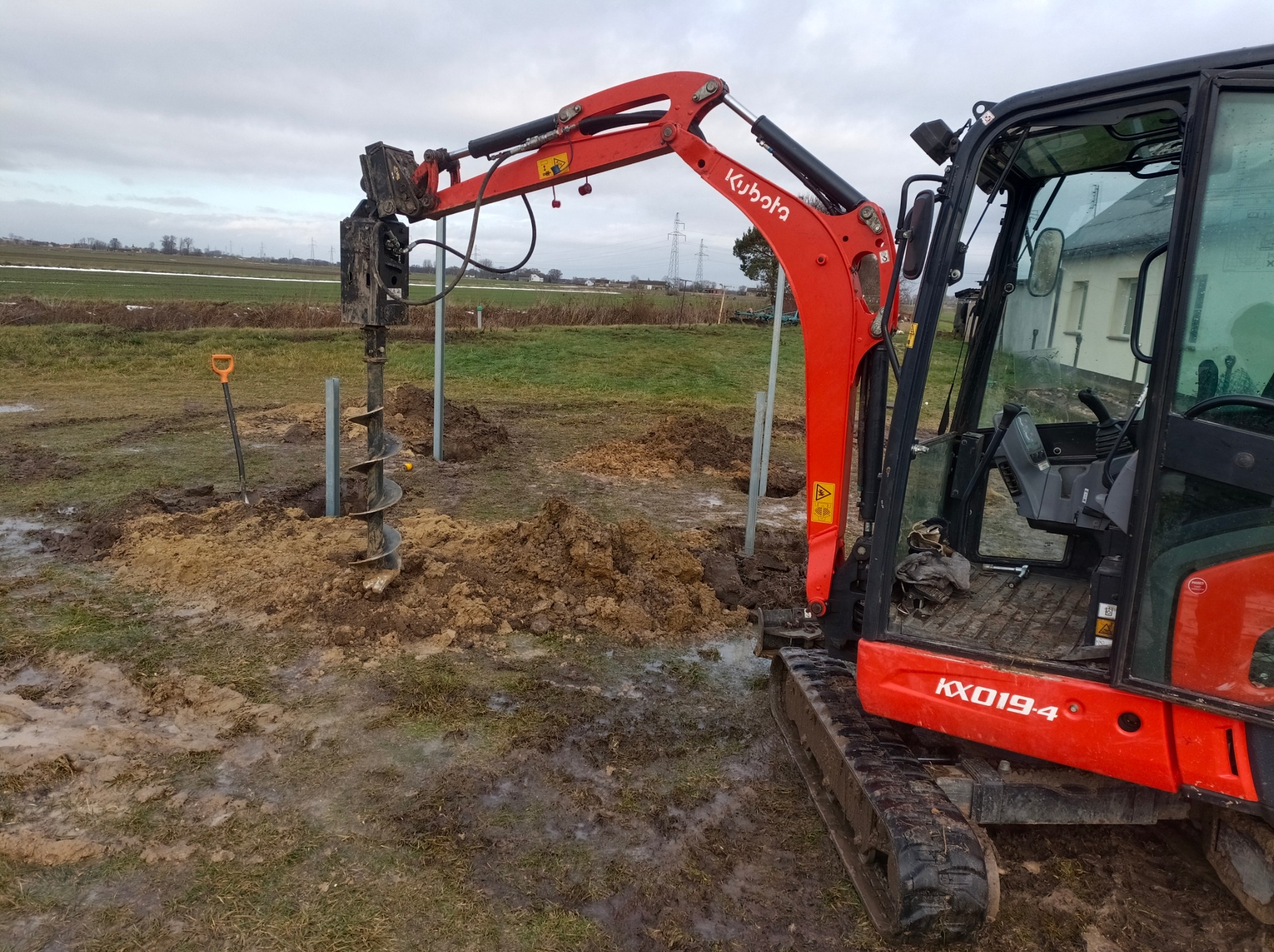 Czerwona minikoparka Kubota KX019-4 z wiertnicą hydrauliczną w trakcie wiercenia otworów w grząskim terenie, widoczne metalowe słupki i łopata w tle.