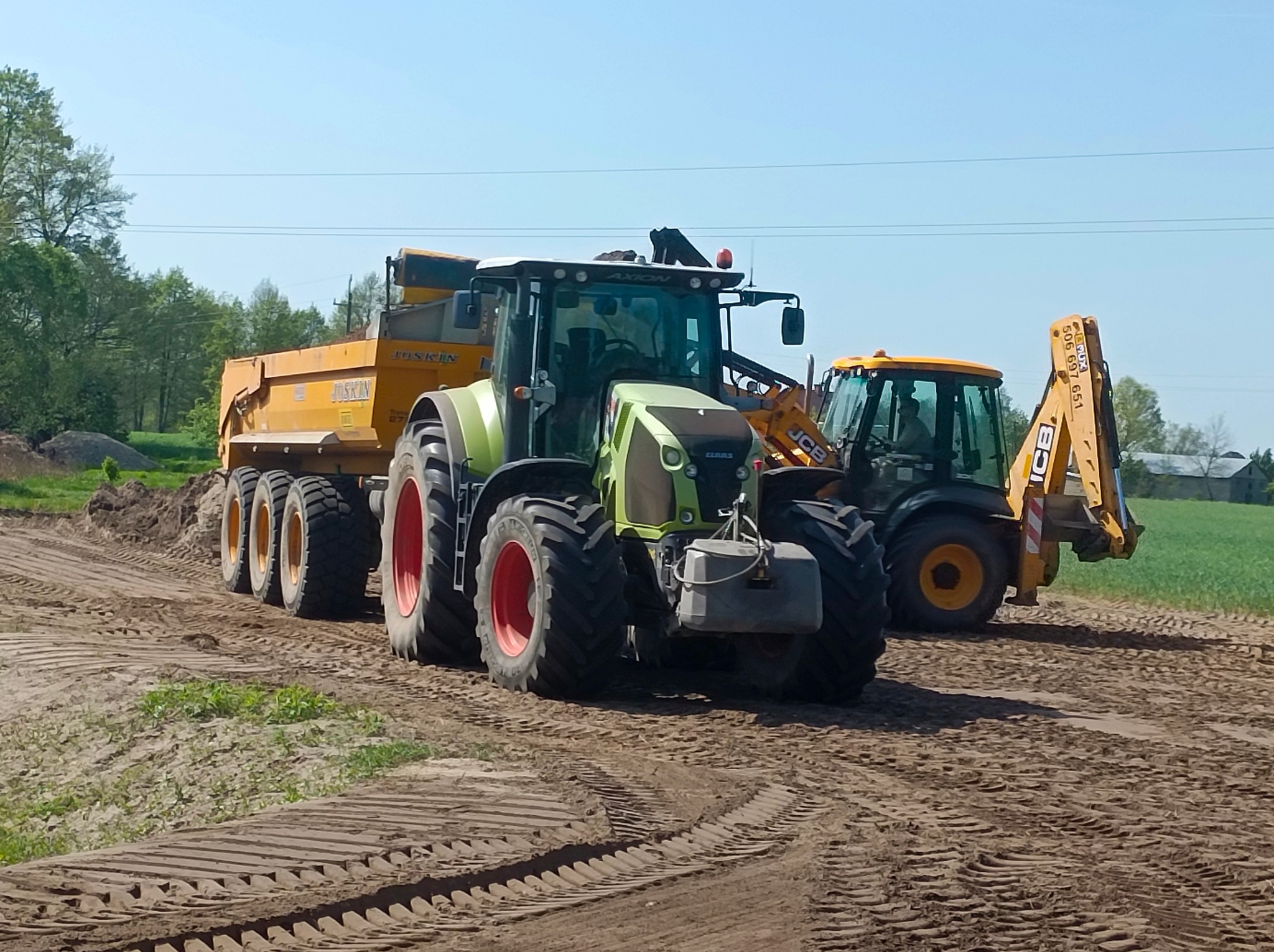 Zielony traktor Claas Axion z żółtą przyczepą Joskin i żółta koparko-ładowarka JCB na polu z widocznymi śladami opon.