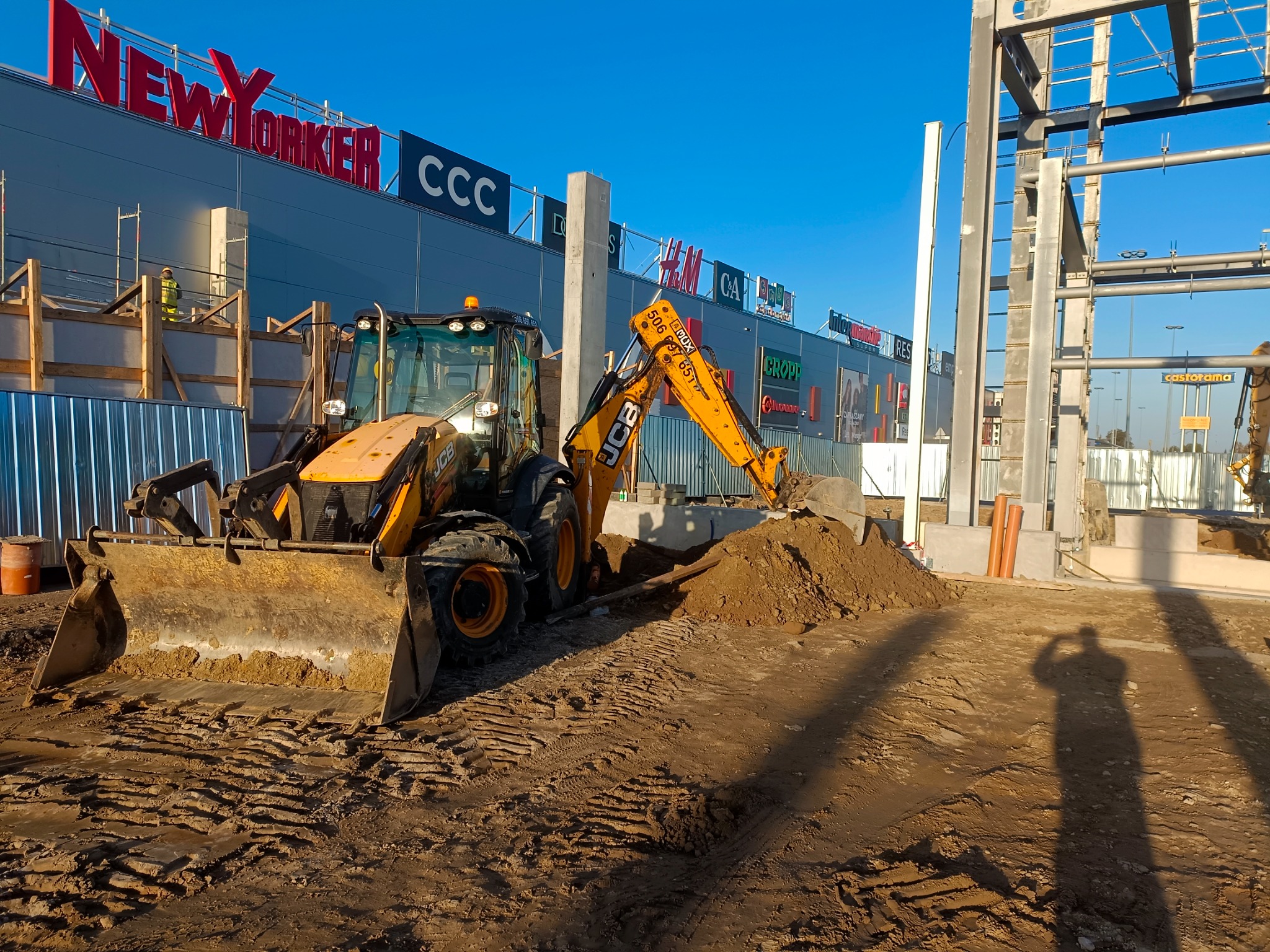 Żółta koparko-ładowarka JCB nabiera ziemię na placu budowy centrum handlowego, widoczne konstrukcje stalowe i fundamenty pod budynki.
