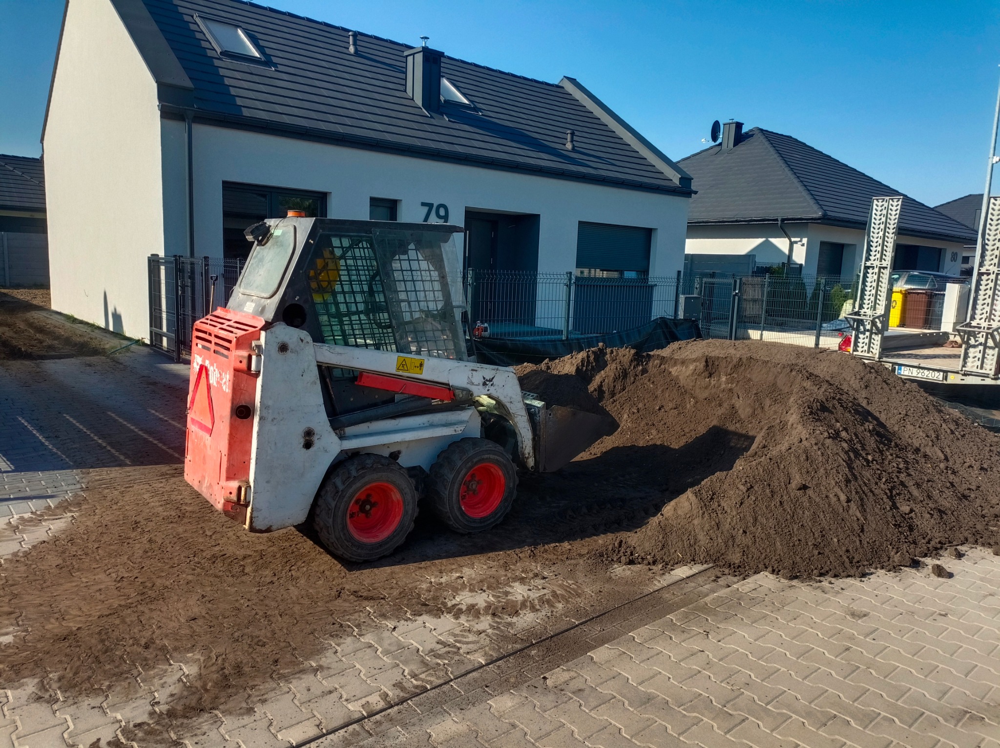 Miniładowarka Bobcat z czerwoną ramą i kołami, usypuje pryzmę ciemnej ziemi na tle nowoczesnych domów jednorodzinnych z ciemnymi dachami.