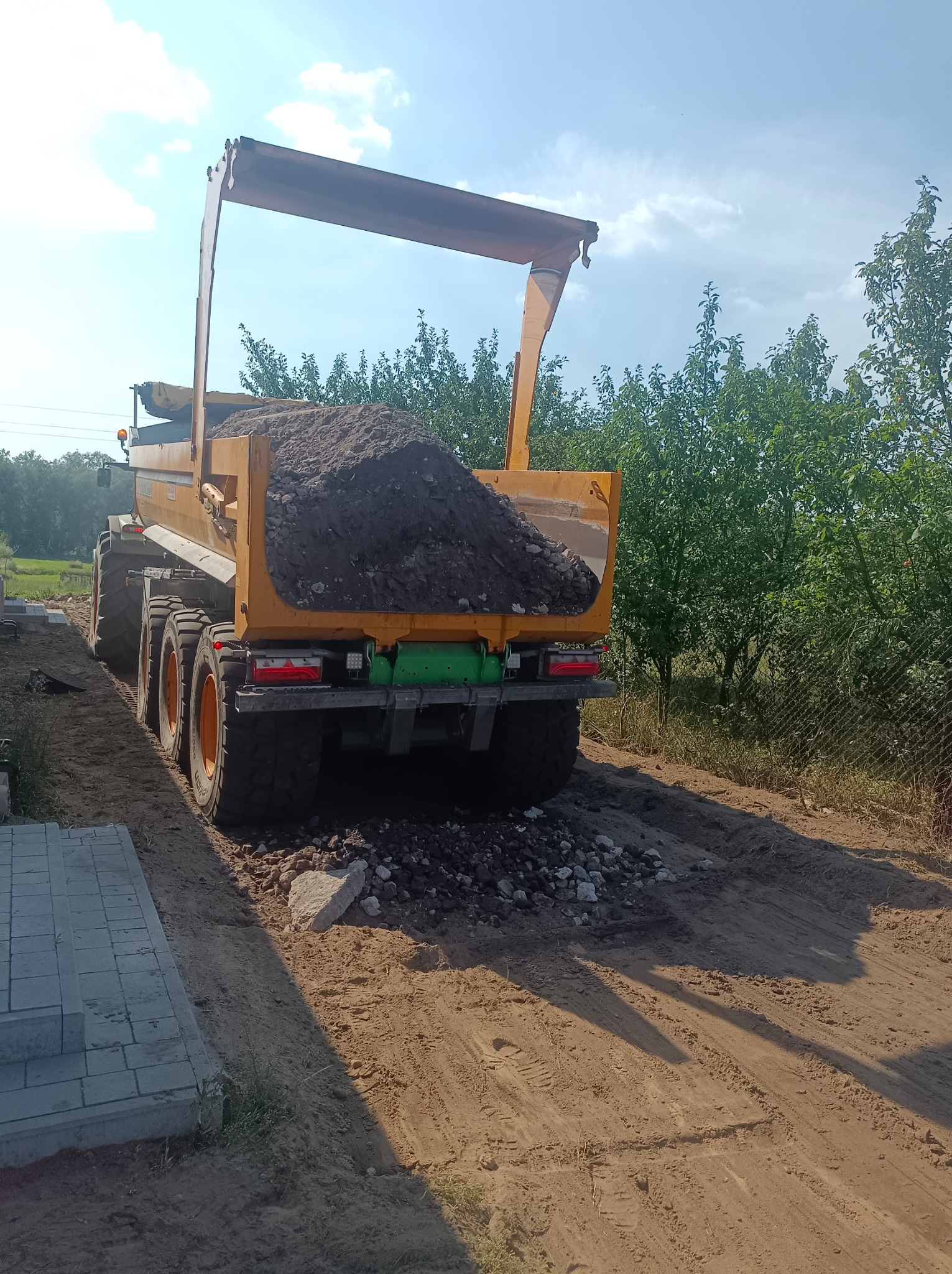 Żółta wywrotka z ładunkiem ciemnej ziemi lub gruzu na nierównej, piaszczystej drodze w słoneczny dzień, widok z tyłu.