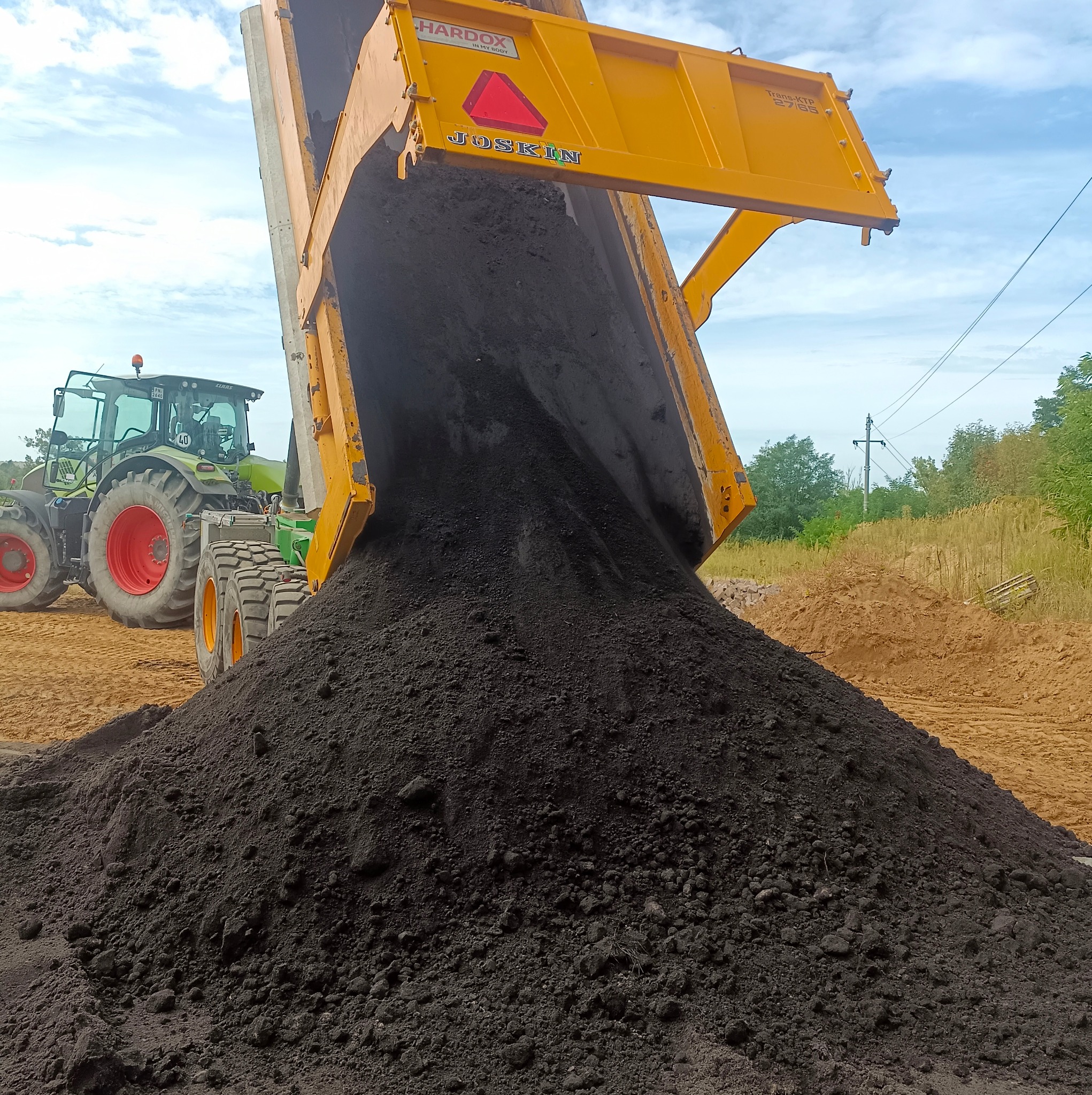 Żółta wywrotka marki Joskin wysypuje ciemną ziemię na plac budowy; w tle widoczny zielony traktor Claas.