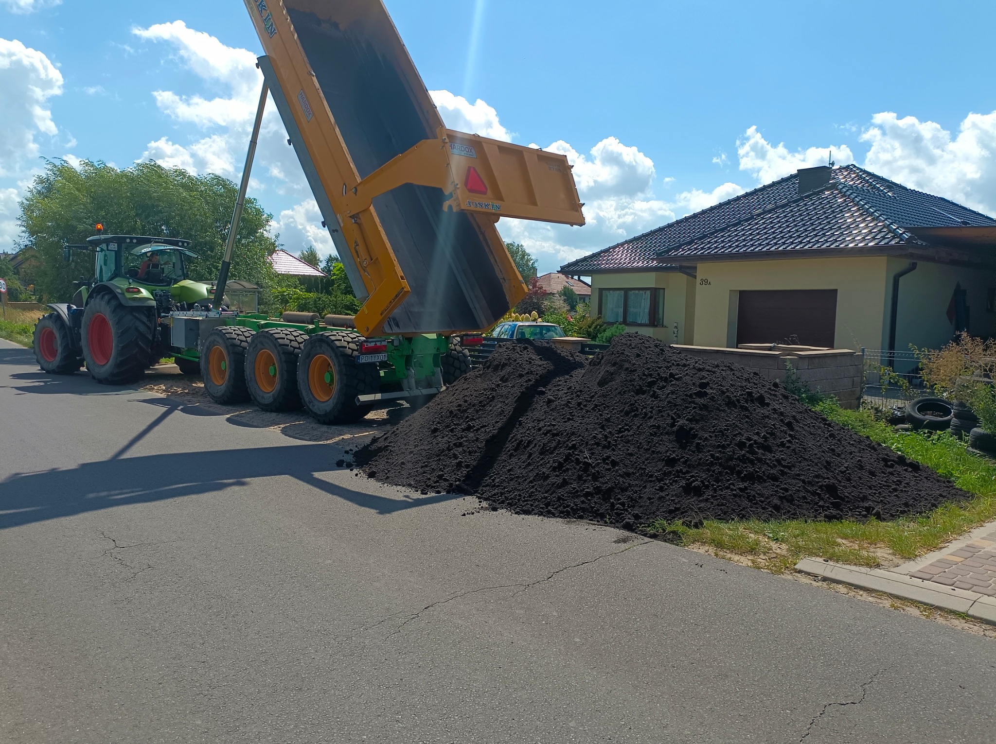 Zielony traktor z żółtą przyczepą wyładowuje ciemną ziemię na poboczu asfaltowej drogi, w tle dom jednorodzinny pod błękitnym niebem.
