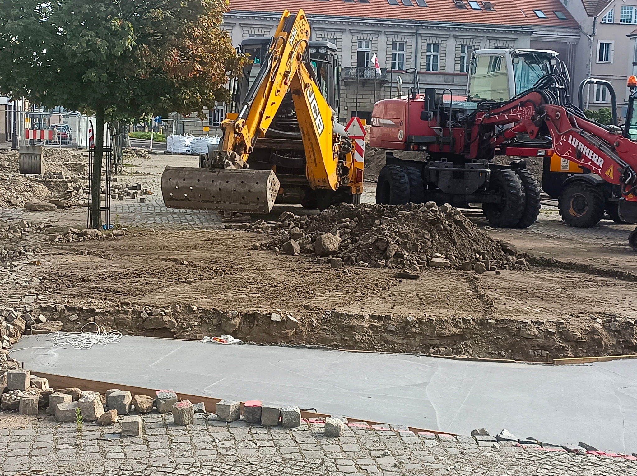 Przygotowanie terenu pod budowę: żółta koparka JCB i czerwona Liebherr na placu budowy, widoczny wykop z betonową wylewką i porozrzucane kostki brukowe, w tle budynki miejskie.