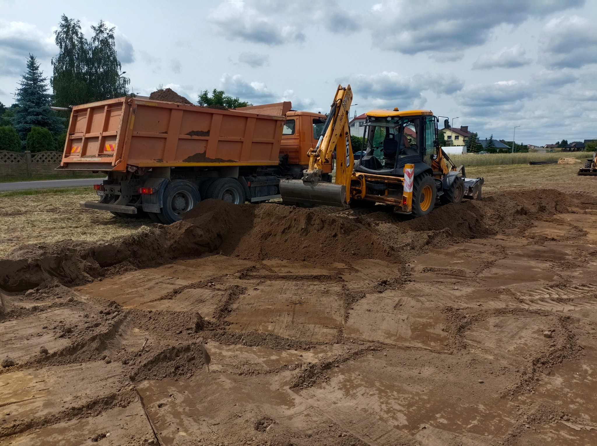 Żółta koparko-ładowarka JCB i pomarańczowa wywrotka podczas prac ziemnych na placu budowy, widoczne ślady opon w ziemi i wykop pod szambo.