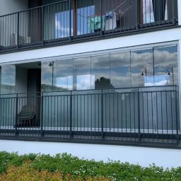 Cover Glass - Nowoczesny balkon z przesuwnymi szklanymi panelami i metalową balustradą, widok z zewnątrz na tle budynku mieszkalnego z zielenią w pierwszym planie.