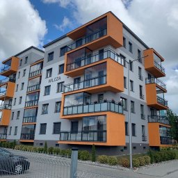 Cover Glass - Nowoczesny budynek mieszkalny z balkonami z balustradami ze szkła, pomalowanymi na pomarańczowo, widok z zewnątrz, na pierwszym planie ogrodzenie.