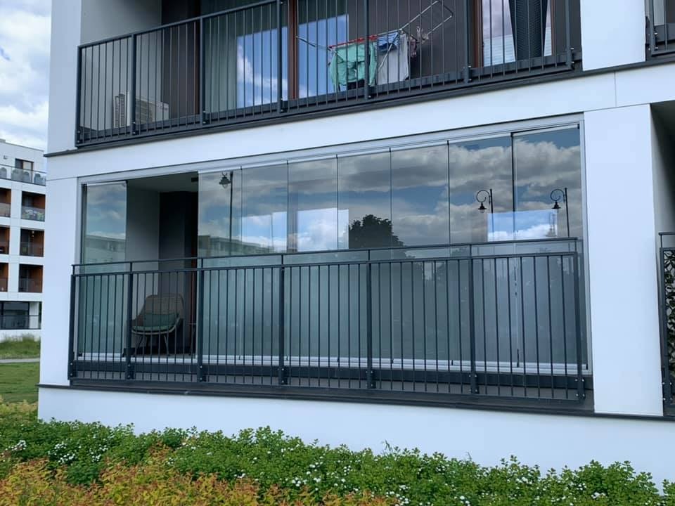 Nowoczesny balkon z przesuwnymi szklanymi panelami i metalową balustradą, widok z zewnątrz na tle budynku mieszkalnego z zielenią w pierwszym planie.