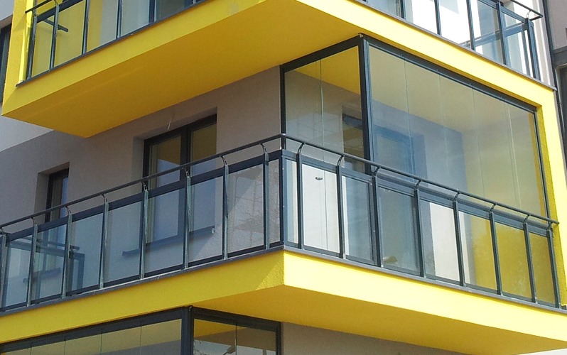 Żółty balkon z czarną, metalową balustradą i szklaną zabudową, widok z dołu.