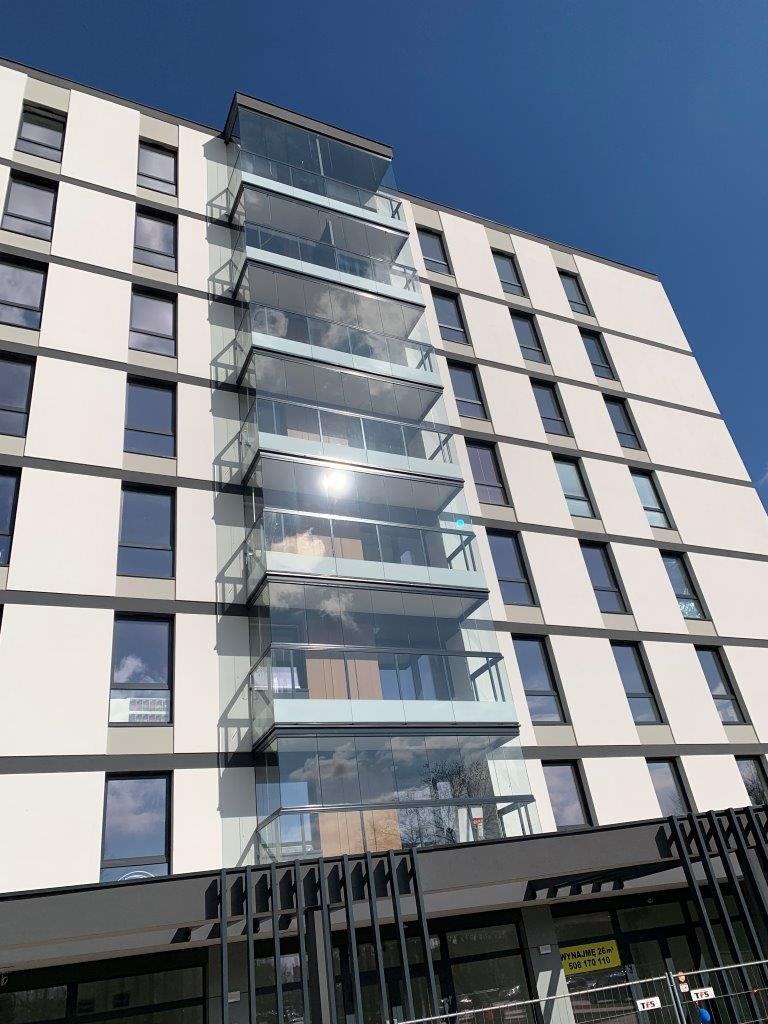 Nowoczesny budynek z przeszklonymi balkonami i balustradami, widok z dołu w słoneczny dzień.