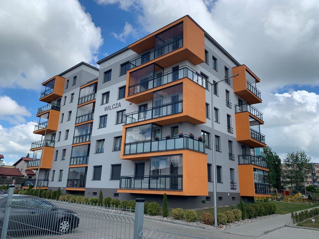 Nowoczesny budynek mieszkalny z balkonami z balustradami ze szkła, pomalowanymi na pomarańczowo, widok z zewnątrz, na pierwszym planie ogrodzenie.