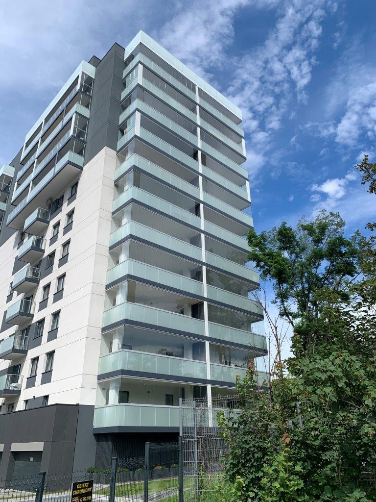 Wysoki budynek mieszkalny z przeszklonymi balkonami, widok z dołu na błękitne niebo z chmurami.